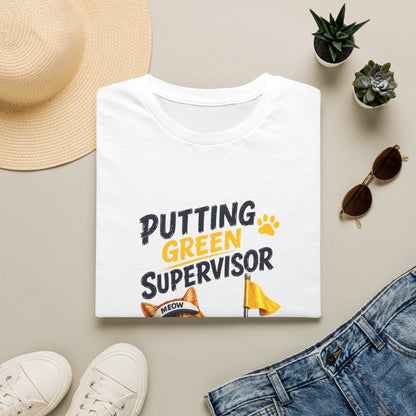 A folded white graphic T-shirt is displayed in a styled flat lay on a light beige surface. The shirt reads 'PUTTING GREEN SUPERVISOR' in black and yellow text with a small cat-and-flag graphic.