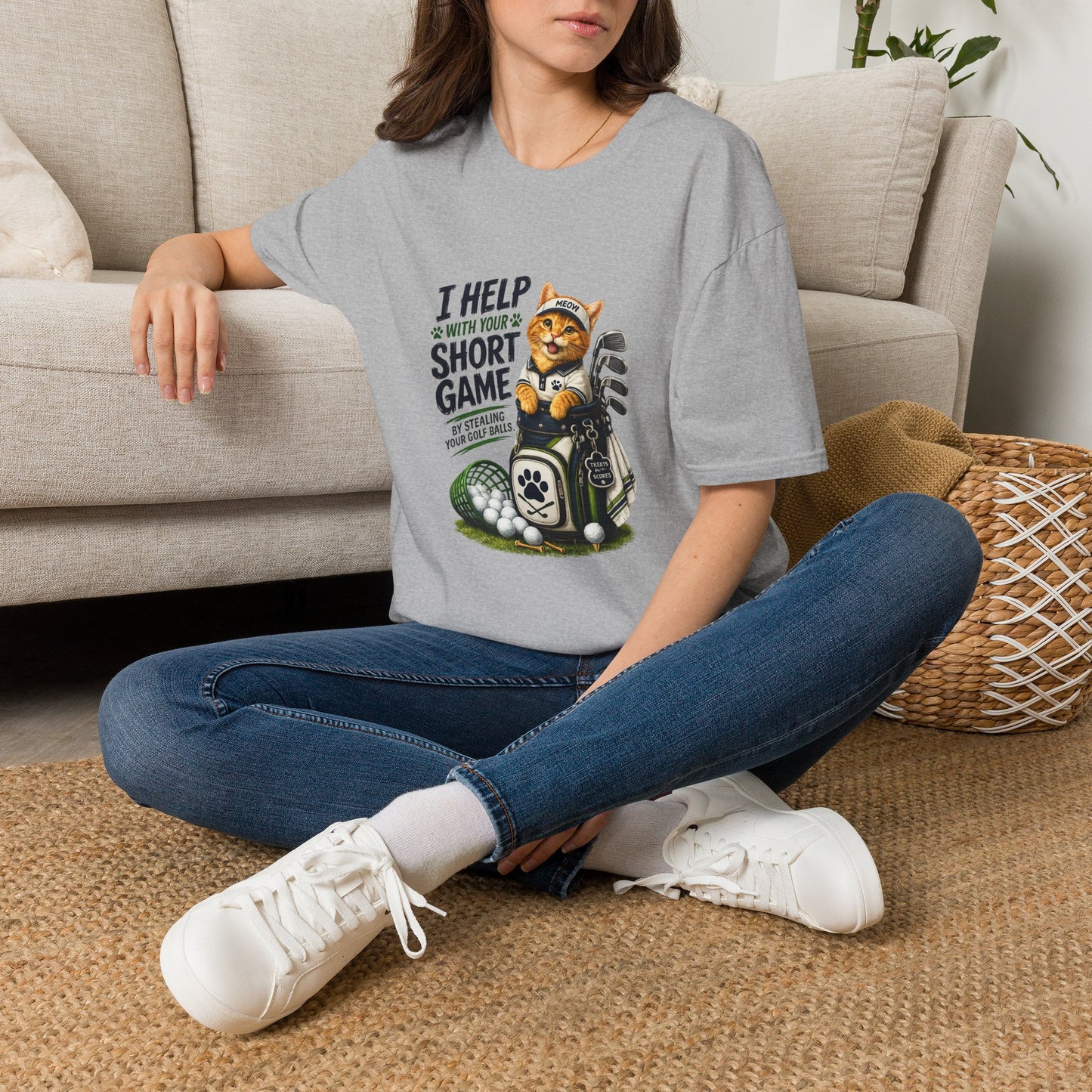 A sport grey graphic T-shirt is displayed in a styled flat lay on a light beige surface. The shirt features a cartoon cat wearing a baseball cap and holding golf clubs, with the text 'I HELP WITH YOUR SHORT GAME BY STEALING GOLF BALLS.