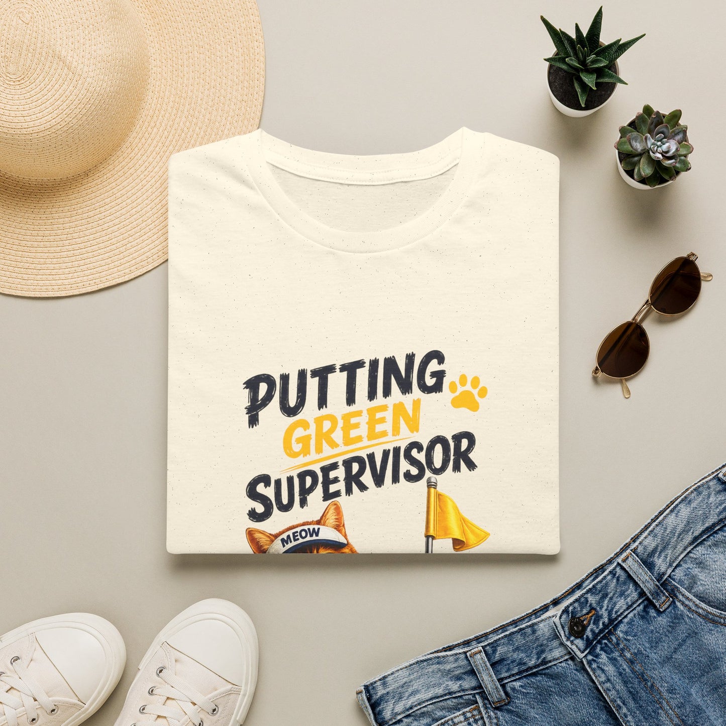 A folded cream-colored t-shirt with a graphic that reads 'PUTTING GREEN SUPERVISOR' in black and yellow, styled in a flat lay with a straw hat, white sneakers, blue jeans, sunglasses, and small potted succulents on a light gray background.