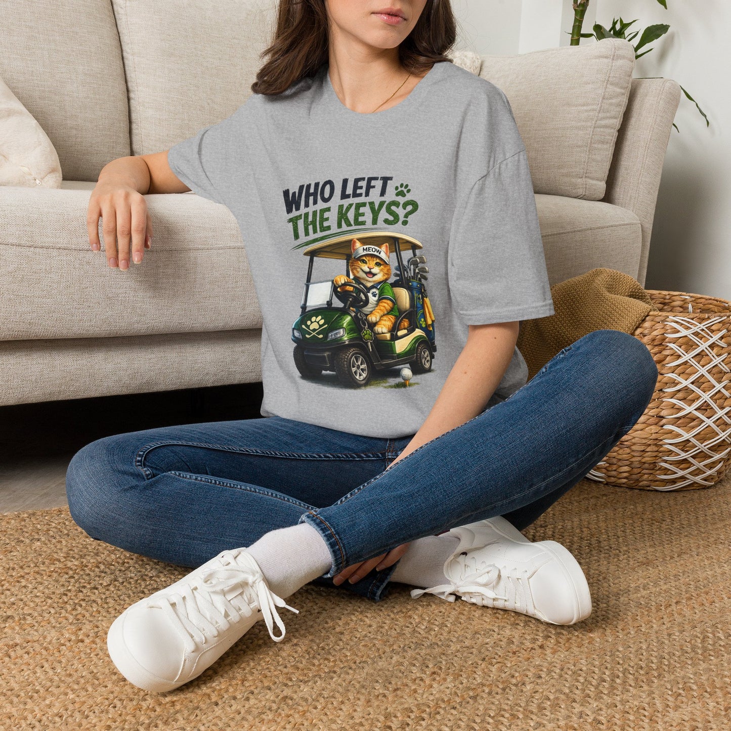 A woman sitting cross-legged on a woven rug in front of a beige sofa, wearing a light gray graphic T-shirt with a golf cart illustration and the text 'WHO LEFT THE KEYS?'.