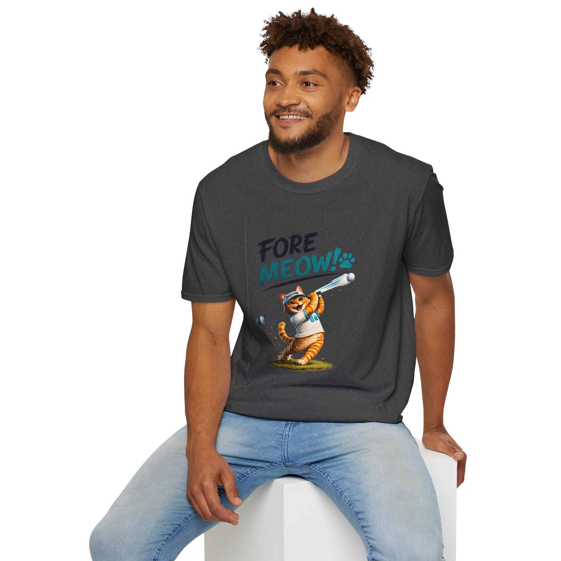 A man wearing a dark gray graphic t-shirt with a cartoon raccoon holding a baseball bat and the text 'FORE MEOW!'. He is sitting on a white block against a plain white background.