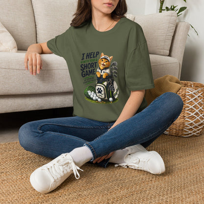 A military green graphic T-shirt is displayed in a styled flat lay on a light beige surface. The shirt features a cartoon cat wearing a baseball cap and holding golf clubs, with the text 'I HELP WITH YOUR SHORT GAME BY STEALING GOLF BALLS.