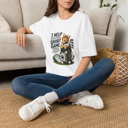 A  white graphic T-shirt is displayed in a styled flat lay on a light beige surface. The shirt features a cartoon cat wearing a baseball cap and holding golf clubs, with the text 'I HELP WITH YOUR SHORT GAME BY STEALING GOLF BALLS.