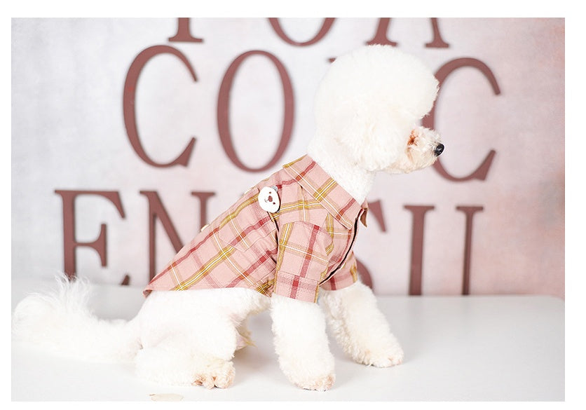 White dog wearing a pink plaid outfit with a button, standing on a white surface with a decorative background.