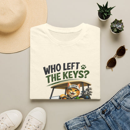 A folded cream-colored t-shirt is displayed in a styled flat-lay arrangement on a light gray surface. The shirt features a graphic print with the text 'WHO LEFT THE KEYS?' in dark green and black, accompanied by a cartoon illustration of an orange tabby cat driving a golf cart.