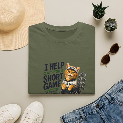 A folded military green graphic T-shirt is displayed in a styled flat lay on a light beige surface. The shirt features a cartoon cat wearing a baseball cap and holding golf clubs, with the text 'I HELP WITH YOUR SHORT GAME BY STEALING GOLF BALLS.