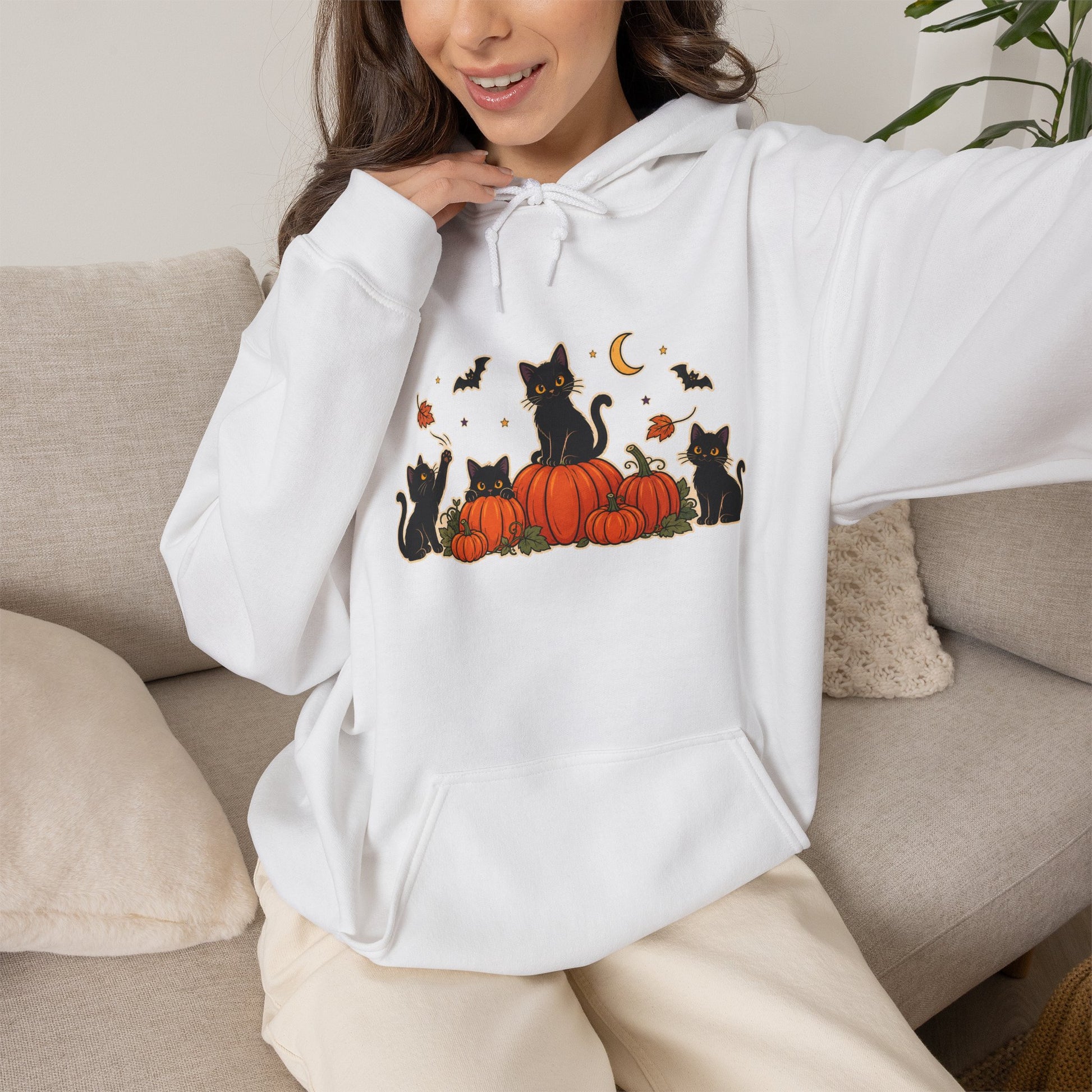 A woman wearing a white pullover hoodie with a graphic of black cats sitting on orange pumpkins, surrounded by small autumn details like bats, leaves, and a crescent moon, posed on a beige couch.