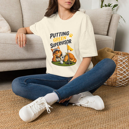 A woman sitting cross-legged on a woven rug in front of a beige sofa, wearing a cream-colored graphic t-shirt with a cartoon cat golf design and the text 'PUTTING GREEN SUPERVISOR'.