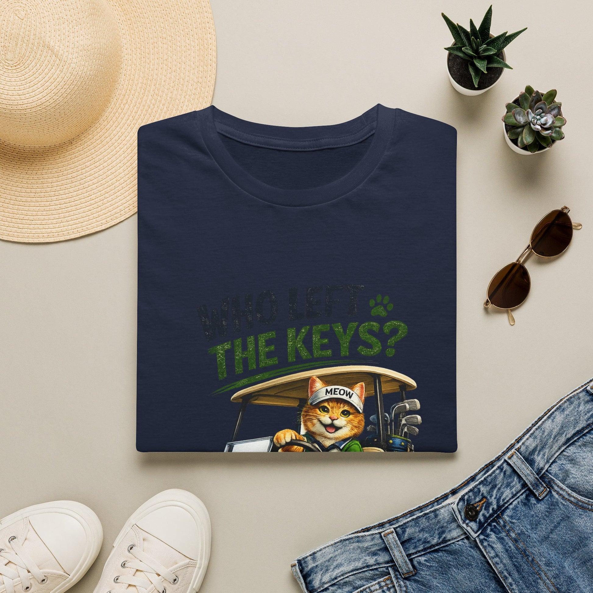 A folded navy graphic T-shirt is displayed in a styled flat lay on a light beige surface. The shirt features a cartoon cat driving a golf cart with the text 'WHO LEFT THE KEYS?' and a paw print.