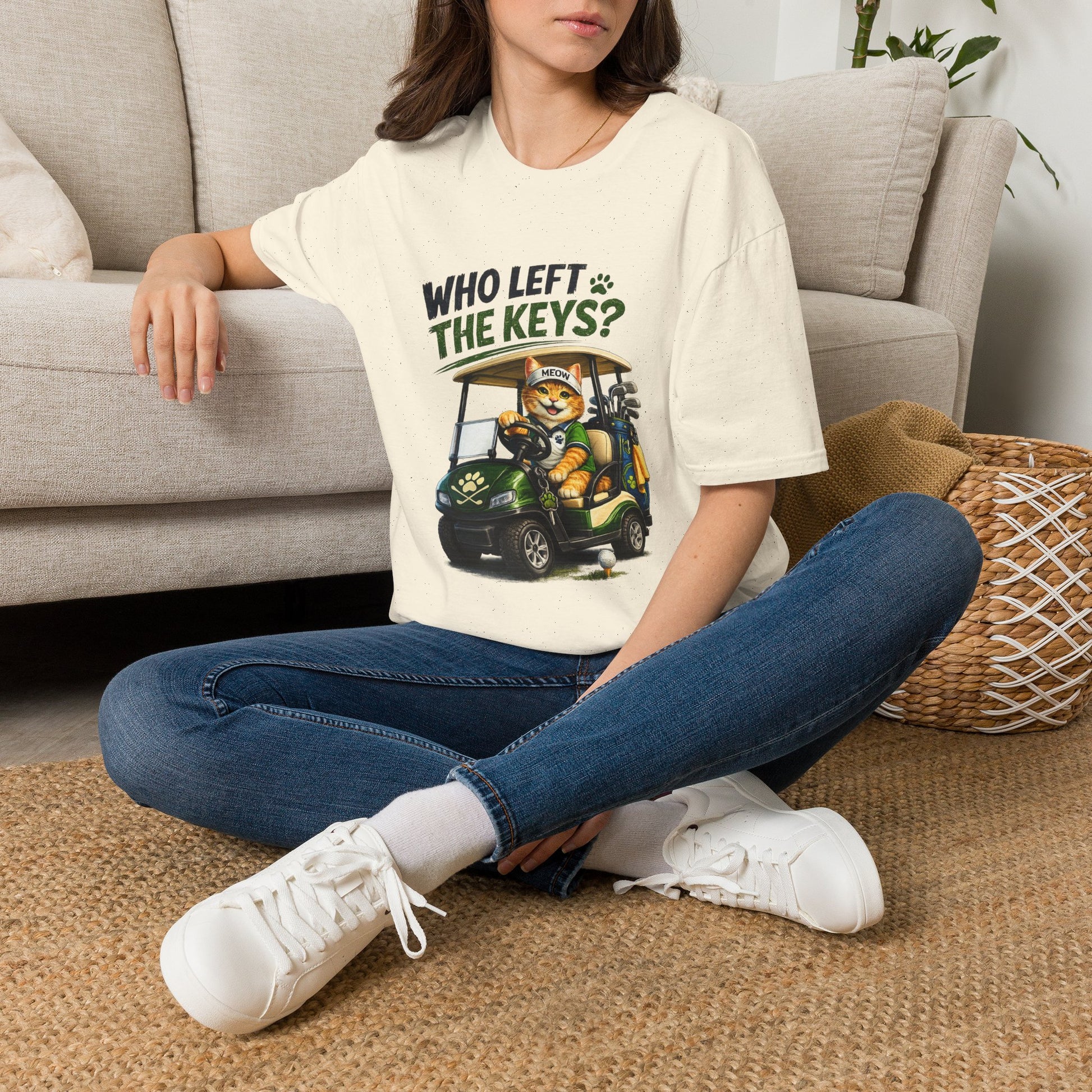 A woman sitting cross-legged on a woven rug in front of a beige sofa, wearing a cream graphic T-shirt with a cartoon cat driving a golf cart and the text 'WHO LEFT THE KEYS?'.