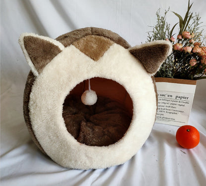 Cat-shaped pet bed with a small ball inside, placed on a white surface with a plant and paper in the background.