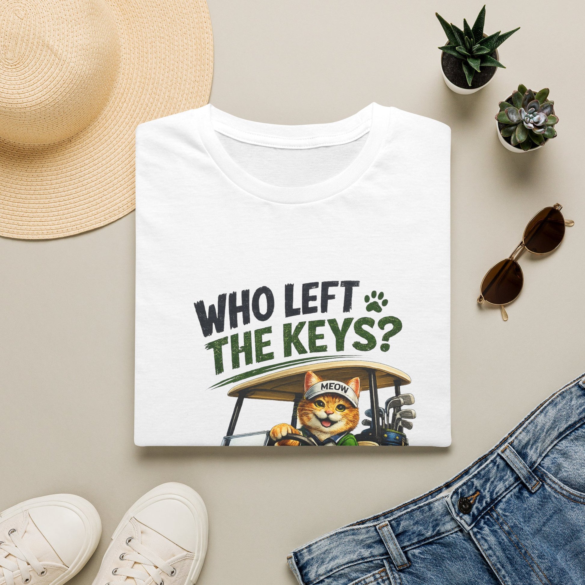 A folded white graphic T-shirt is displayed in a styled flat lay on a light beige surface. The shirt features the text 'WHO LEFT THE KEYS?' in dark green and black, with a cartoon cat driving a golf cart.