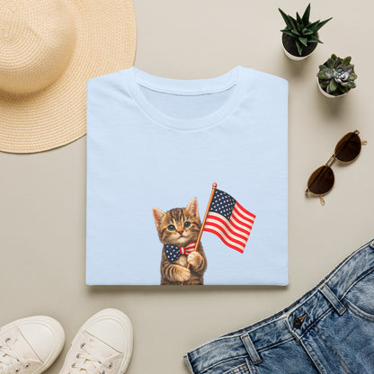 Light blue t-shirt with a kitten holding an American flag design, surrounded by a hat, sunglasses, shoes, and plants on a beige surface.