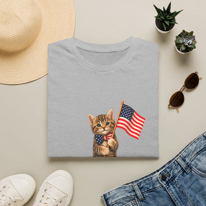 Gray t-shirt with a cat holding an American flag design, surrounded by a hat, sunglasses, shoes, and plants on a light surface.