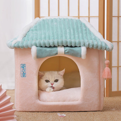 Cat peeking out from a pink and teal pet house with tassels on a light wooden floor.