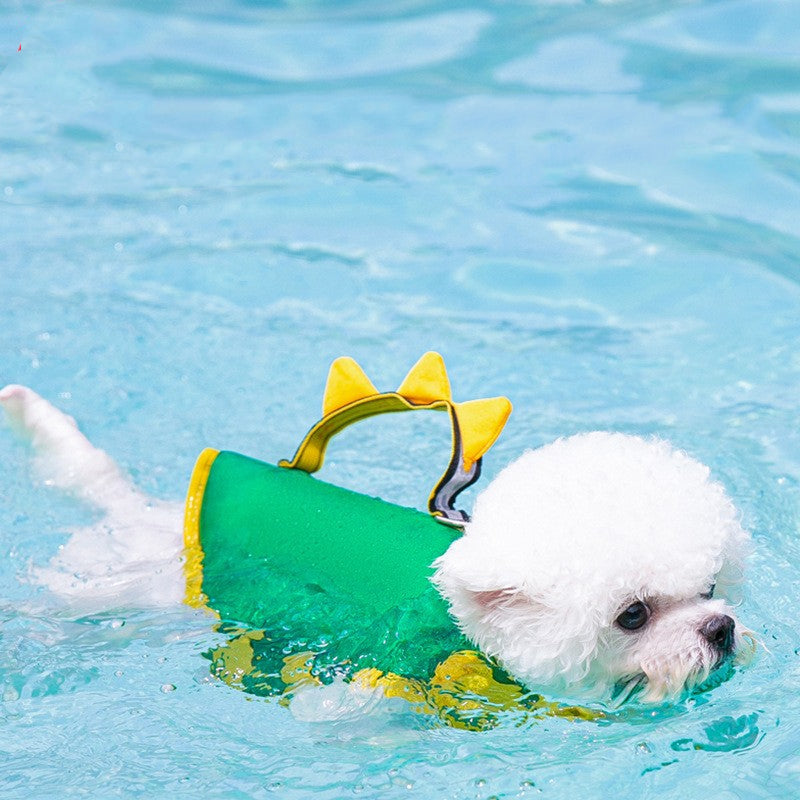 Small white dog wearing a green and yellow swim vest with shark fin design, swimming in clear blue water.