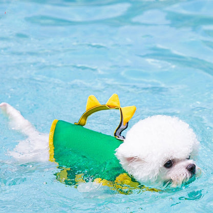 Small white dog wearing a green and yellow swim vest with shark fin design, swimming in clear blue water.